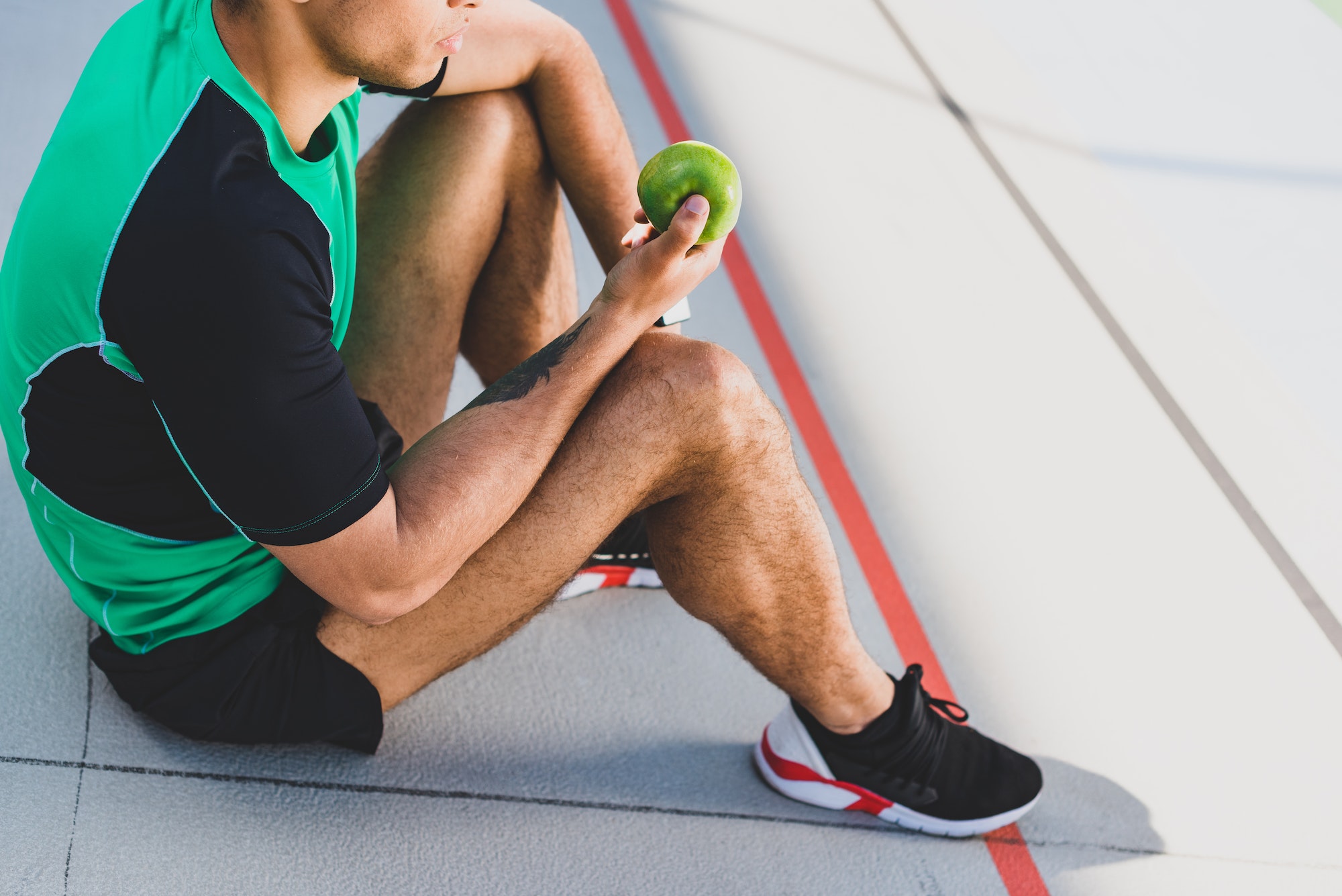 cropped view of sportsman sitting on running track and holding green apple